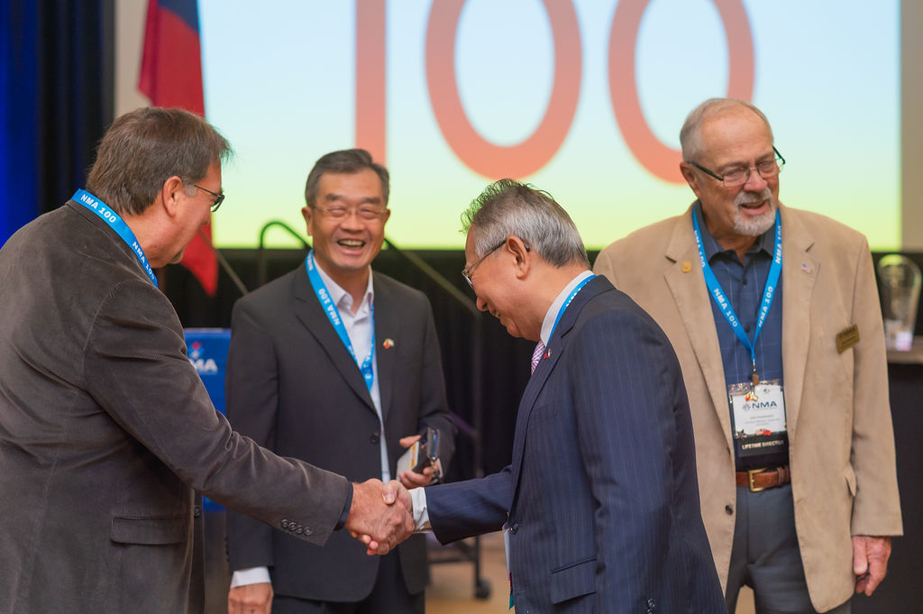 Conference attendees shaking hands in front of an NMA 100 anniversary screen